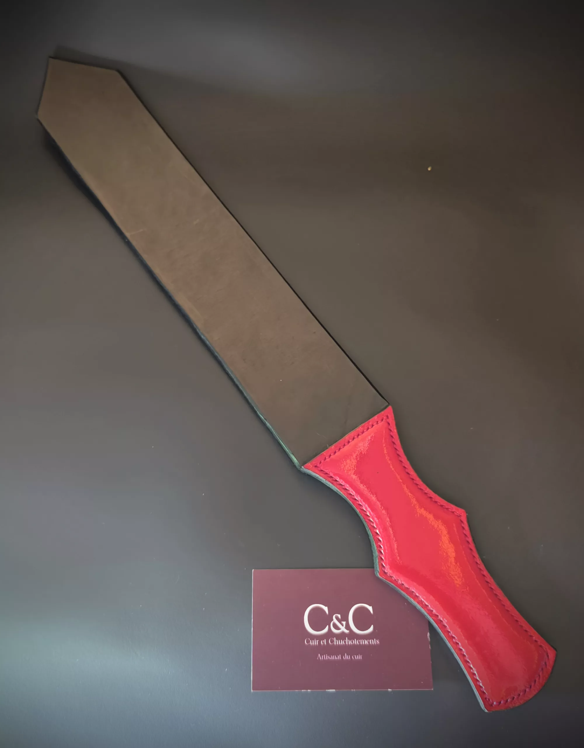 Red leather strap with burgundy edge lying on a dark surface, partially covering a maroon card that reads 'C&C Cuir et Chouk...' (brand label visible)