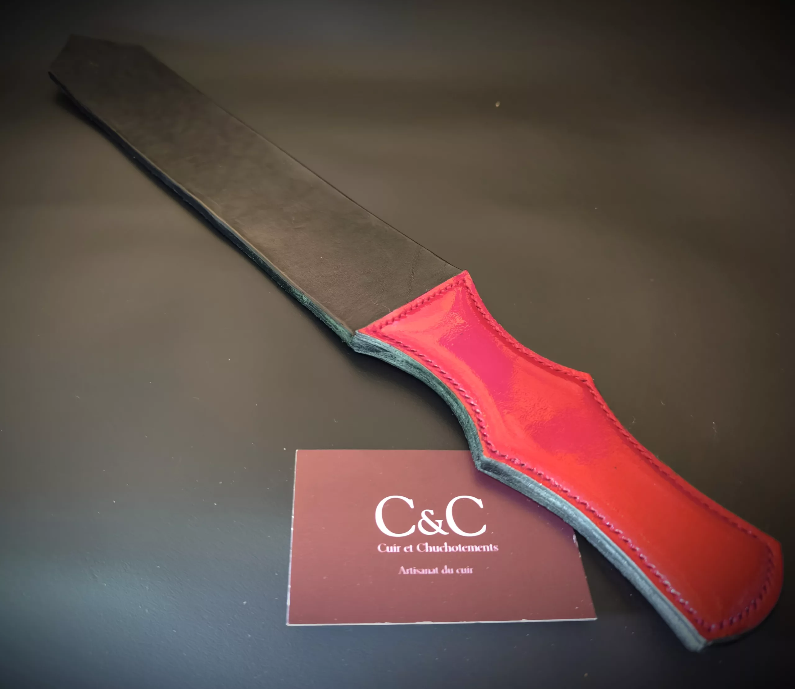 Close-up of a red leather strap with teal edge on a dark surface, a burgundy card with 'C&C' partially visible nearby.